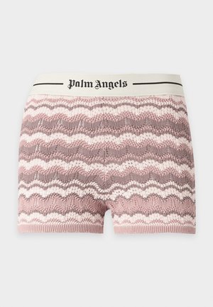 Mladá žena stojaca s rukou na bok, nosi svetloružové tričko Palm Angels, vzorované pletené šortky a čierne baleríny na svetlom pozadí.