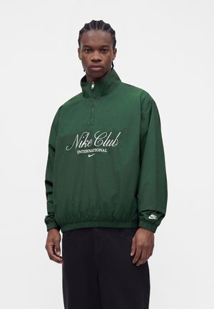 Ung mand med flettet hår iført en mørkegrøn Nike Club-pullover med hvidt logo og sorte bukser mod en ensfarvet baggrund.