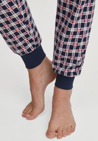 Blote voeten met een gestructureerd navyblauw, wit en rood geruit pyjamabroek met rode strikjes, tegen een effen lichte achtergrond.
