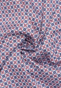 Katoenen stof met een herhalend geometrisch patroon van rode bloemen en blauwe contouren op een witte achtergrond, met een gestructureerde afwerking.