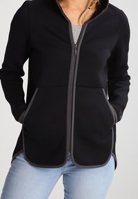 Giacca nera con zip realizzata in tessuto morbido, dotata di cappuccio, due tasche frontali con bordi grigi e orlo arrotondato. Indossata sopra un maglione grigio.