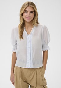 Blouse blanche en voile transparent avec manches bouffantes, bordure en dentelle festonnée le long du col et de la patte de boutonnage, portée sur un haut clair ; associée à un pantalon beige.