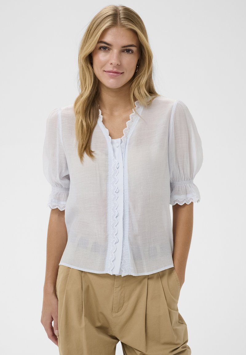 Blouse blanche en voile transparent avec manches bouffantes, bordure en dentelle festonnée le long du col et de la patte de boutonnage, portée sur un haut clair ; associée à un pantalon beige.