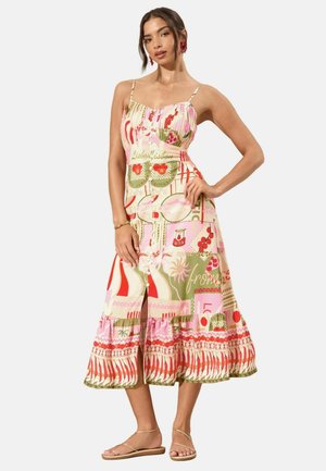 Frau trägt ein bunt gemustertes Midikleid mit Spaghettiträgern, Knopfleiste vorne und Rüschensaum, kombiniert mit flachen Sandalen mit Riemchen.