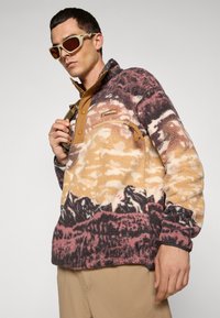 Man met beige zonnebril en een multikleuren fleece trui met bergpatronen en beige broek tegen een effen achtergrond.