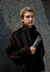 Veste polaire noire avec accents rouges, col montant ; personne tenant un tube de crème coiffante gris étiqueté "CURL CREAM". Fond gris texturé.