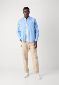 Lichtblauw geruit overhemd met lange mouwen, gedragen over een wit T-shirt. Beige loszittende broek en witte sneakers.