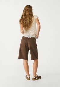 Mujer con pelo largo y ondulado, vistiendo una blusa corta de mangas de encaje blanca, pantalones cortos marrones hasta la rodilla y zapatillas marrones con suelas blancas.