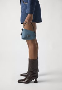 Bolsa de denim em miniatura com detalhes pretos, apresentando uma forma triangular e um logótipo dourado. Usada com botas de couro castanho brilhante até ao joelho.