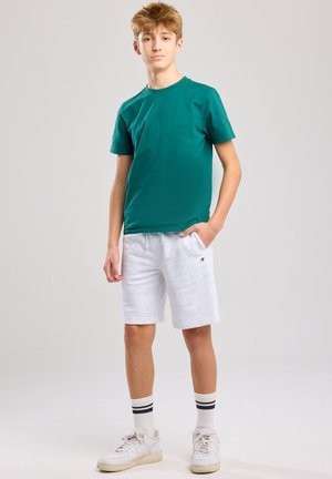 Grünes Rundhals-T-Shirt, weiße Baumwollshorts mit Taschen und weiße Sneaker mit beigen Akzenten. Gestreifte Socken runden das Outfit ab.