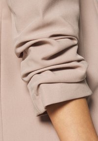 Tissu rose clair côtelé avec des poignets repliés, mettant en valeur une texture lisse et un subtil éclat. Gros plan sur le bras portant le vêtement.