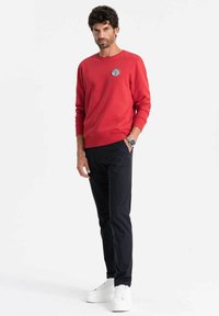 Sudadera roja de mezcla de algodón con cuello redondo y parche de logo negro, combinada con pantalones oscuros y zapatillas blancas. Fondo liso.