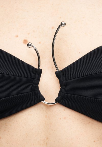 Haut de bikini noir côtelé maintenu par une fermeture métallique incurvée argentée avec des extrémités en forme de boule, sur une poitrine à peau claire parsemée de taches de rousseur.