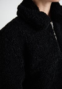 Close-up of a person wearing a black textured fleece jacket with a high collar and visible silver zipper.