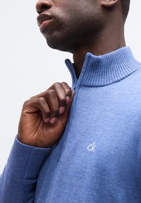 Κοντινό πλάνο ενός ατόμου που κλείνει το φερμουάρ ενός ανοιχτό μπλε ριγωτού πουλόβερ με γιακά, στο στήθος του οποίου υπάρχει μικρό λευκό λογότυπο «ck».