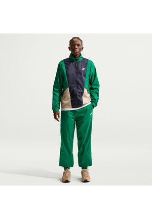Person steht und trägt einen grünen und marineblauen Nike Trainingsanzug mit beige-farbenen Akzenten und beigen Sneakers vor einem schlichten hellen Hintergrund.