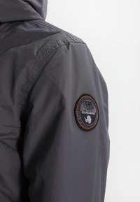 Grå jakke med en glat, skinnende tekstur og et rundt sort og orange logoemblem på venstre ærme. Har synlige syninger.