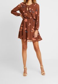 Brun blomtryckt klänning med långa ärmar, nyckelhålsringning, elastiskt midjeband och flytande kjol. Accenterad med rosa och blå blommor.