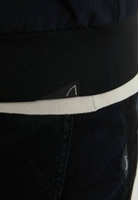 Gros plan sur une ceinture côtelée noire posée sur un tissu crème et un pantalon sombre avec de petites étiquettes de vêtement noires triangulaires attachées.