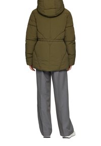 Olijfgroene pufferjack met een capuchon, voorzien van stikdetails en een taille met aansnoering. Gepaarde met grijze wijde broek en witte sneakers.