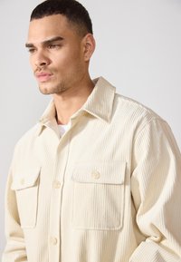Chemise en velours côtelé de couleur crème avec un col, une fermeture à boutons sur le devant et deux poches poitrine. Présente des côtelés verticaux et une coupe décontractée.