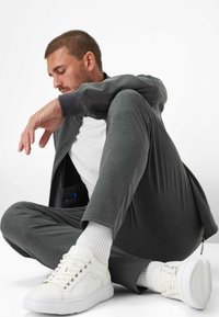 Grijze sportjack en bijpassende broek, gecombineerd met witte sneakers. De broek heeft een ritsdetail en het jack heeft een zwarte zak.