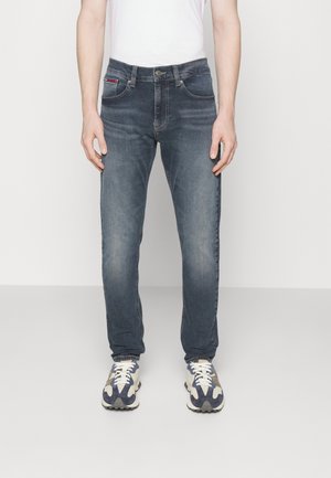 Mann trägt ausgewaschene blaue Jeans mit geradem Bein, weiße Sneaker mit blauen und beigen Akzenten und ein weißes, eingestecktes Hemd, steht auf einem einfachen Boden.