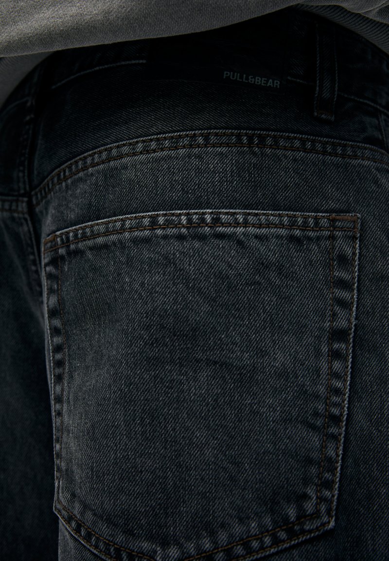 Jeans in denim nero con una superficie testurizzata, caratterizzati da cuciture a contrasto e un'etichetta del logo visibile sulla tasca posteriore.
