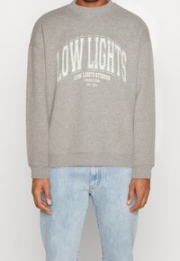 Sudadera gris con el texto "LOW LIGHTS" en letras blancas grandes, corte relajado, cuello redondo, puños acanalados, combinada con vaqueros azules.