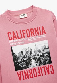 Różowy sweter z napisem "CALIFORNIA" w pogrubionym czerwonym kolorze, z czarno-białym zdjęciem miejskiego pejzażu poniżej, przedstawiającym wieżowce i ulice.