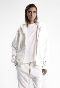 Veste à capuche blanche à fermeture éclair en tissu doux, avec poches avant et cordon de serrage, superposée à un t-shirt blanc uni, associée à un pantalon de jogging assorti.