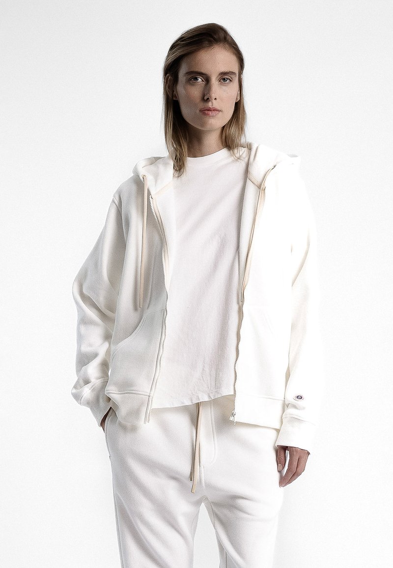 Veste à capuche blanche à fermeture éclair en tissu doux, avec poches avant et cordon de serrage, superposée à un t-shirt blanc uni, associée à un pantalon de jogging assorti.