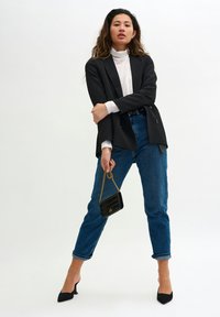 Donna che indossa un blazer nero, un dolcevita bianco, jeans blu arrotolati, tacchi neri, mentre tiene una piccola borsa nera con catena dorata su uno sfondo bianco.