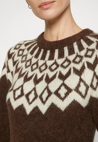 Pull en tricot marron avec un motif géométrique en losanges blancs et marron, doté d'un col rond et d'une texture douce.