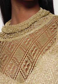 Gros plan d'une personne à la peau foncée portant un haut en maille métallique dorée avec un col haut et un motif ajouré en forme de losanges.