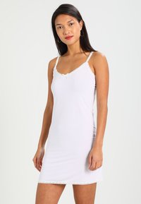 Abito slip in satin bianco con bordo di pizzo lungo il collo e l'orlo. Spalline sottili e texture liscia mettono in risalto un design semplice ed elegante.