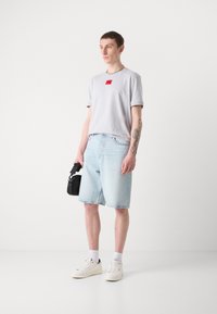 T-shirt grigia chiara con etichetta rossa, pantaloni corti in denim blu e sneaker bianche. Gli accessori includono una piccola borsa nera. Texture lisce, vestibilità casual.