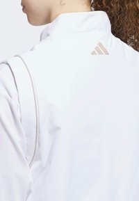Hvid atletisk jakke med høj krave, der har et tekstureret stof og et lysebeige Adidas-logo på ryggen.