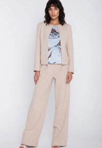 Kobieta stoi, ubrana w beżowe spodnie o szerokich nogawkach, dopasowaną marynarkę z odkrytym przodem oraz niebieską bluzkę w wzory na jednolitym białym tle.