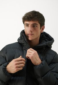 Black puffer jacket with a high collar, zippered front, and elastic cuffs. Smooth texture, filled for insulation, worn by a model.