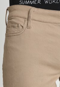 Béžové bavlněné kalhoty s texturovaným povrchem, které mají přední kapsu a šitý pásek s nápisem "SUMMER WORLD" v bílé barvě.