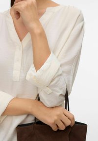 Personne portant une chemise blanche à manches longues retroussées, tenant les poignées d'un sac à main en daim marron foncé sur un fond uni.