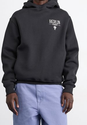 Osoba ubrana w czarną bluzę z kapturem z napisem "BERLIN GERMANY" oraz grafiką balonu na gorące powietrze, w jasnoniebieskich spodniach, stojąca na białym tle.