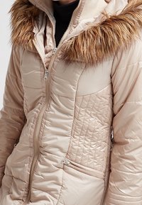 Giacca puffer beige chiaro con cappuccio bordato in pelliccia, caratterizzata da una texture trapuntata, tasche laterali e una chiusura con zip completa.