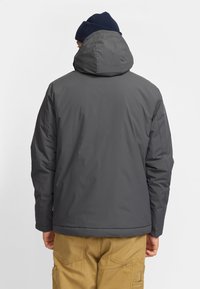 Chaqueta acolchada gris con un alto cuello y capucha incorporada, con textura suave. Combinada con pantalones cargo color tostado, que muestran bolsillos a los lados.