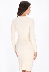 Knie-lange, geribde gebreide jurk in crème kleur met lange mouwen en een aansluitende silhouet. Beschikt over een ronde halslijn en minimale details.