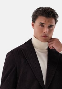 Dunkelbrauner Cord-Blazer mit dicken vertikalen Streifen und einem scharfen Revers. Darunter kontrastiert ein cremfarbener Rollkragenpullover mit dem strukturierten Stoff.