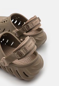 Brown synthetic sandals with an enclosed heel and adjustable fabric strap. Features ventilation holes and textured sole for grip.