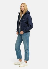 Giubbotto bomber navy con zip frontale, cappuccio regolabile e polsini a costine, abbinato a jeans azzurri chiari e sneakers bianche.