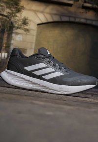 Baskets Adidas noir et blanc avec semelle Cloudfoam, trois bandes blanches sur le côté, posées sur un trottoir urbain avec un arrière-plan flou.